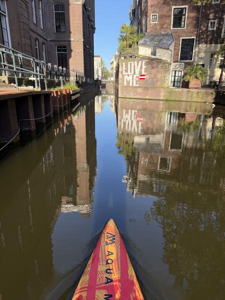 The photo captures a serene view from a paddleboard gliding along the Grimburgwal canal in Centrum, Amsterdam, under a bright blue sky